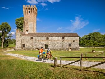 Zwei Radfahrer auf einem Schotterweg vor dem Castello di San Martino, einer mittelalterlichen Steinfestung mit hohem Turm unter blauem Himmel.