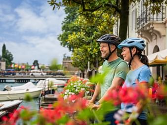 Zwei lächelnde Radfahrer mit Helmen stehen an einem blumengesäumten Kanal in Peschiera am Gardasee, mit Booten und historischen Gebäuden im Hintergrund.