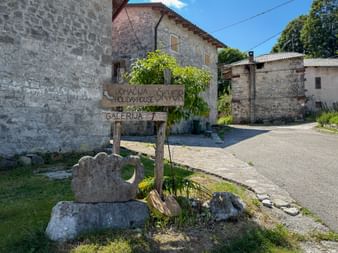 Wooden signpost with text 'Domačija Škvor Holiday House Galerija' in front of stone buildings in Robidišče village on a sunny day.