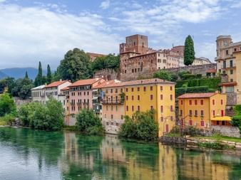 Bunte historische Gebäude entlang des Flusses Brenta in Bassano del Grappa mit mittelalterlichem Turm und Burg auf dem Hügel.