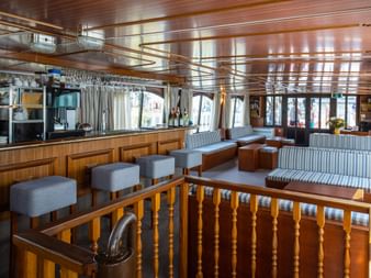 Innenbereich des Schiffes MS Lestello mit Holzbar, hängenden Gläsern, gestreiften Sitzbänken und großen Fenstern mit weißen Vorhängen.