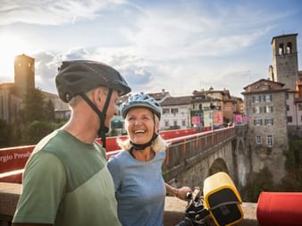 Smiling couple with helmets on a red bridge in Cividale del Friuli, with historic buildings and a bell tower in the background.