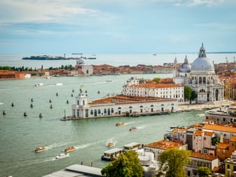 Luftaufnahme der Lagune von Venedig mit Booten, historischen Gebäuden mit Terrakottadächern und weißer Kuppelkirche. Ferne Inseln am Horizont.