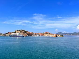 View of Piombino harbor with cruise ship and boats in turquoise water. Historic town and fortress visible on hillside under blue sky.