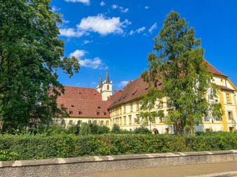 Gelbe historische Gebäude mit roten Ziegeldächern und Kirchturm der Gösser Brauerei unter blauem Himmel, eingerahmt von grünen Bäumen.