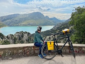 Radfahrer sitzt auf Steinmauer am Cuber-Stausee im Tramuntana-Gebirge, Mallorca. Türkisfarbenes Wasser und Berggipfel im Hintergrund.