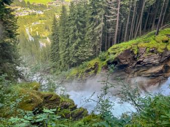 Krimmler Wasserfall stürzt über moosbewachsene Felsen durch dichten Wald. Nebel steigt vom fallenden Wasser auf mit grüner Vegetation im Vordergrund.