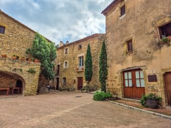 Historische Steinhäuser umgeben einen Kopfsteinpflasterhof in Katalonien. Zypressen und Topfpflanzen schmücken den Platz mit Holztüren und Fenstern.