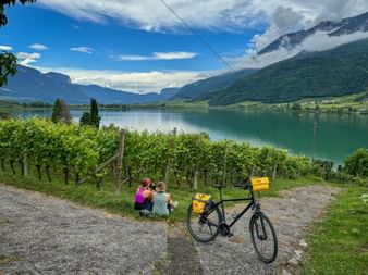 Zwei Radfahrer sitzen in einem Weingarten mit Blick auf den Kalterer See in Südtirol. Ein Tourenrad mit gelben Taschen steht daneben, Berge sind über dem See sichtbar.