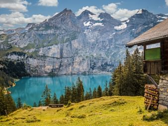 Holzhütte auf grüner Wiese mit Blick auf türkisen Alpensee im Berner Oberland. Schneebedeckte Berggipfel und Wälder im Hintergrund.