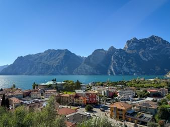 Panoramic view of Torbole on Lake Garda with colorful buildings, turquoise water, and dramatic mountain peaks under clear blue sky.