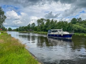 Blue and white motor yacht cruising on a calm canal near Ossenzijl. Green grass banks and trees line the waterway under a cloudy sky.
