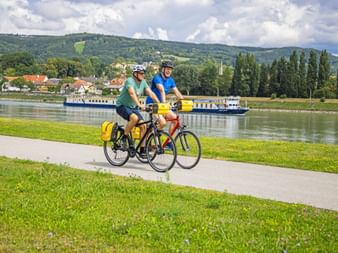 Zwei Radfahrer auf Uferweg entlang der Donau mit Flussschiff im Hintergrund, grüne Hügel und Dorf auf der anderen Seite sichtbar.