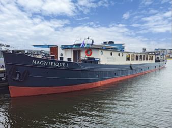 MS Magnifique I river cruise ship moored in harbor. Dark blue hull with red bottom, white upper deck with windows, under blue sky.