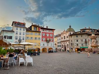 Piazza Duomo in Trient with colorful historic buildings, outdoor café seating, and a fountain. Evening sky with clouds above the square.