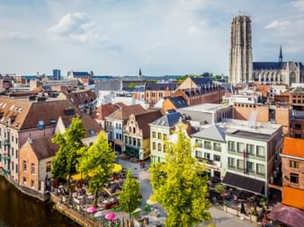 Luftaufnahme des Mechelner Stadtzentrums mit historischen Gebäuden, baumbestandenem Platz und dem hohen Turm der St.-Rombouts-Kathedrale.