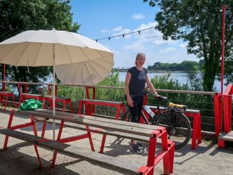 Female cyclist with bike standing at red picnic tables with umbrella on Loire riverside terrace near Nantes, with river and trees in background.