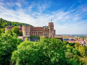 Ruine des Heidelberger Schlosses auf einem Hügel umgeben von grünen Bäumen, mit Blick auf die Stadt und das Tal unter blauem Himmel.
