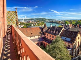 Panoramic view from Basel Minster showing colorful tower roof, historic buildings, Rhine River, and distant hills under blue sky.
