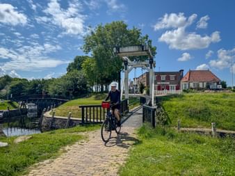 Cyclist crossing a small drawbridge in Vollenhove with canal, green embankments, and traditional Dutch houses under blue sky with clouds.