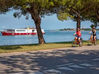 Zwei Radfahrer auf orangen Rädern fahren am Uferweg auf Lido di Venezia mit rot-weißem Boot und Venedig-Skyline im Hintergrund unter blauem Himmel.