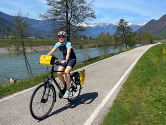 Radfahrerin mit gelben Packtaschen auf dem Etschradweg. Schneebedeckte Berge im Hintergrund, grüne Wiesen und Bäume säumen die Route.