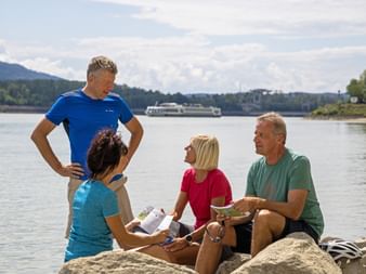Four cyclists reviewing travel documents on rocks by the Danube River. A river cruise ship is visible in the background on the water.