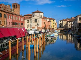 Canale Vena in Chioggia mit hölzernen Pfählen, Booten und bunten Häusern. Eine Kirche mit rotem Ziegeldach und Glockenturm ist sichtbar.