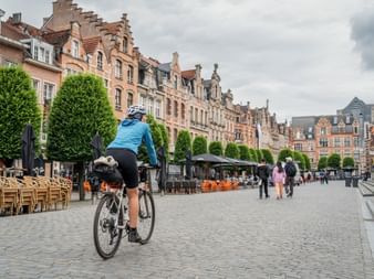 Radfahrer im blauen Trikot auf dem Kopfsteinpflaster des Oude Markt in Leuven, Flandern, mit historischen Treppengiebelhäusern und Cafés.