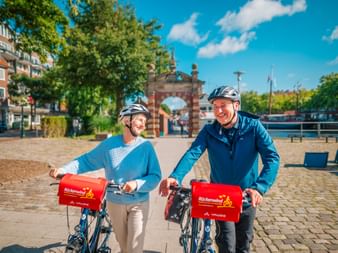Zwei lächelnde Radfahrer mit Helmen und roten Vaude-Taschen auf Kopfsteinpflasterplatz. Historisches Tor und Uferpromenade im Hintergrund unter blauem Himmel.