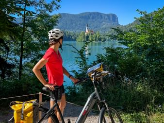 Radfahrerin im roten Shirt mit Fahrrad blickt auf Bleder See mit Inselkirche und Bergen. Gelbe Packtaschen am Rad, umrahmt von grünen Bäumen.