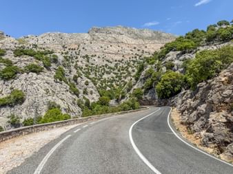 Kurvenreiche Bergstraße in Mallorcas Tramuntana mit Felsklippen und grüner Vegetation unter blauem Himmel.