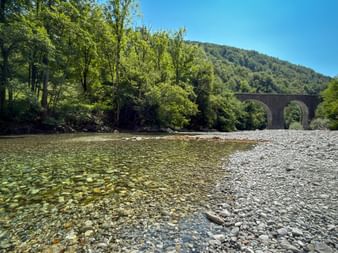 Clear Bača River with pebble beach and stone viaduct bridge in background, surrounded by green forested hills under blue sky near Tolmin.