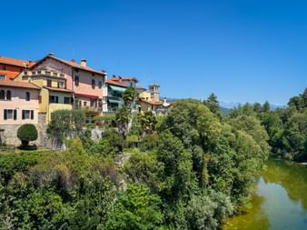 Colorful houses of Cividale del Friuli perched on a cliff above a green river, surrounded by lush vegetation under a clear blue sky.