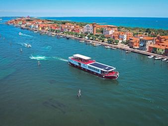 Aerial view of MS Ave Maria cruise ship in blue-green water near Pellestrina with colorful waterfront buildings and wooden pier posts.
