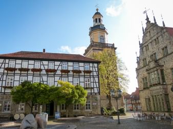 Marktplatz in Rinteln mit Fachwerkhaus, Kirchturm mit gelber Fassade und Uhr, verziertem Renaissance-Rathaus und grünen Bäumen.