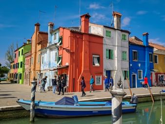 Bunt bemalte Häuser in Rot, Weiß, Blau und Grün säumen einen Kanal in Burano. Ein blaues Boot liegt im Vordergrund, Touristen gehen am Ufer entlang.