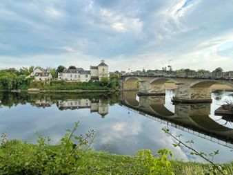 Mehrbogige Steinbrücke über die Loire in Chinon, mit historischen Gebäuden, die sich im ruhigen Wasser spiegeln, und grüner Vegetation im Vordergrund.