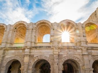 Antike Steinbögen des römischen Amphitheaters in Arles mit Sonnenlicht, das durch die obere Arkade gegen blauen Himmel scheint.