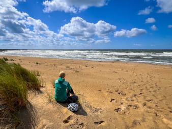 Radfahrer in türkisfarbener Jacke sitzt am sandigen Ostseestrand mit Helm daneben und blickt auf Wellen unter blauem Himmel mit weißen Wolken.