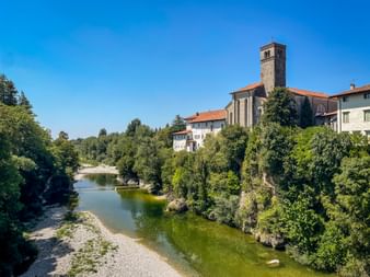 Historic church with bell tower in Cividale del Friuli overlooking the green Natisone River, surrounded by lush trees under blue sky.