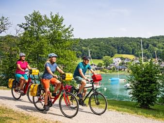 Three cyclists with yellow panniers on a lakeside path at Mattsee. Sailboats on turquoise water, village and green hills in background.