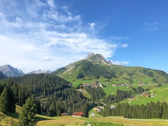 Panoramic view of an Alpine village nestled in green hills with traditional houses scattered across meadows, surrounded by forested mountains.