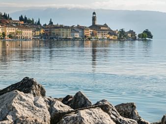 View of Salò town on Lake Garda with historic buildings and church tower reflected in calm water. Rocky shoreline in foreground, mountains in background.