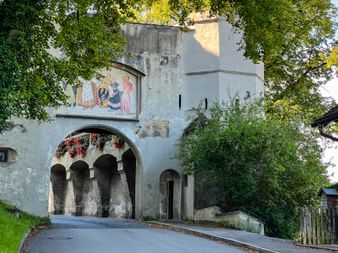 Medieval gate tower in Schongau with white walls, arched passageway, and colorful religious fresco. Red flowers hang from the upper level.
