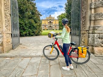 Female cyclist with red bike and yellow panniers viewing Villa Torrigiani through an ornate stone gate in Capannori, Tuscany.