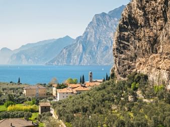 Dorf Torbole mit Kirchturm zwischen Olivenhainen und dramatischen Felsen, mit Blick auf den Gardasee und Bergen im Hintergrund.