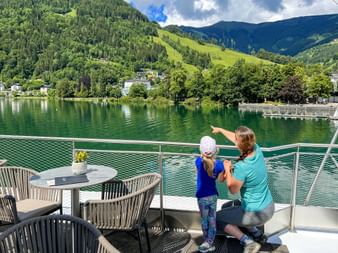 Mother and child on a terrace overlooking Lake Zell with green mountains and forested slopes in the background under a partly cloudy sky.