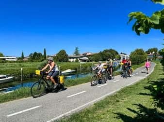 Group of cyclists riding along a paved path beside a canal near Aquileia. Green grass and trees line the route under a clear blue sky.