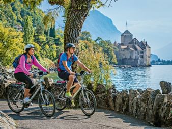 Zwei Radfahrer fahren entlang des Genfersee-Ufers mit Schloss Chillon am Wasser. Berge und Bäume rahmen den malerischen Seeweg ein.